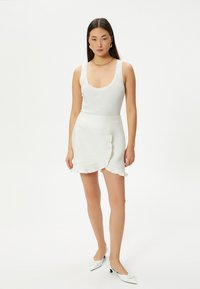 Débardeur blanc à côtes associé à une jupe longue à taille haute avec un ourlet volanté. Le mannequin porte des chaussures blanches à talons pointus et des accessoires dorés simples.