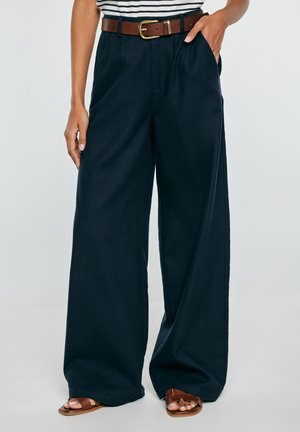 Osoba nosząca granatowe spodnie o wysokim stanie i szerokich nogawkach z brązowym paskiem oraz brązowe sandały z odkrytymi palcami, ręka w kieszeni, częściowo widoczna koszulka w paski.