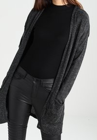 Długi, otwarty kardigan w kolorze ciemnoszarym, wykonany z dzianiny o miękkiej fakturze. Posiada boczne kieszenie oraz luźne, zwiewne rękawy. W zestawieniu z czarną dopasowaną bluzką i spodniami.