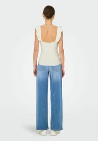 Ribbet, off-white ærmeløs top med flæse-stropper og åben ryg, parret med lyseblå vide jeans og hvide sneakers.