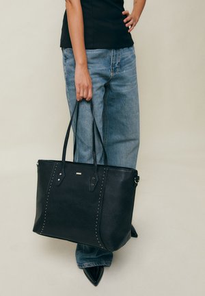 Person iført blå jeans og sort top holder en stor sort lædertaske med nitte detaljer og lange remme.