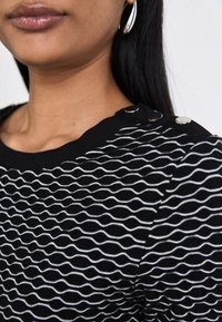 Kobieta ubrana w czarno-białą bluzkę w faliste paski z metalowymi guzikami na ramieniu i widocznymi srebrnymi kolczykami w kształcie kółek, w pobliżu długich, czarnych włosów.