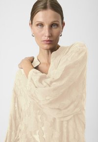 Šviesiai béžinė nėrinių bluzonė su gėlių raštu, ilgomis rankovėmis ir laisvu siluetu. Savybė - kuklus iškirpimas ir švelni tekstūra.