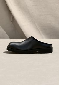 Μαύρο δερμάτινο mule παπούτσι τύπου slip-on με στρογγυλεμένη μύτη και χαμηλό τακούνι σε ανοιχτό μπεζ επιφάνεια και φόντο.