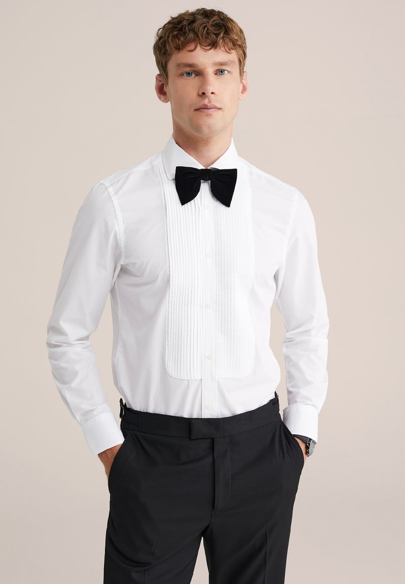 Balta smokings krekls ar pleķētu priekšpusi, melnu tauriņu un garām piedurknēm ar pogām. Valkāts ar melnām biksēm.