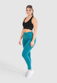 Zwarte sport-bh, teal hoge taille legging en witte sneakers. Gladde textuur met een aansluitend ontwerp en minimale merkopdruk op de tailleband.