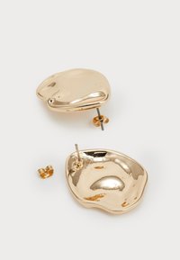 Χρυσές μεταλλικές σκουλαρίκια με κυματιστό, άνισο σχήμα. Έχουν κούμπωμα με στήριγμα και κυκλικό κομμάτι στην πίσω πλευρά για ασφαλή στερέωση.