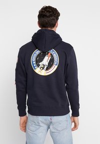 Felpa blu navy con cappa, caratterizzata da una grafica di un navetta spaziale e la scritta "100esima Missione dello Space Shuttle" sul retro, dotata di cappuccio con coulisse.