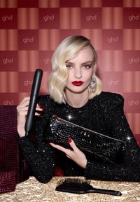Ein schwarzer Haarglätter, eine funkelnde schwarze Clutch und eine schwarze Paddle-Bürste auf einer strukturierten Oberfläche, vor einem gemusterten rot-pinken Hintergrund.