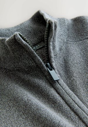 Lähikuva osittain vetoketjulla varustetusta harmaasta neulepaidasta, jossa on resorikaulus ja musta vetoketjun vetolaite.