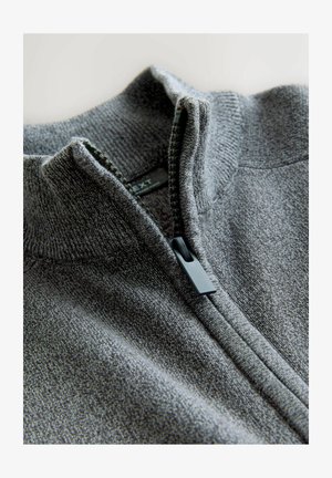 Close-up van een deels geopend grijs gebreid vest met een geribde kraag en een zwarte ritsluiting.