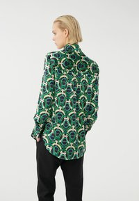 Langarmhemd mit einem geometrischen Muster in Grün, Schwarz und Creme. Hergestellt aus glattem Stoff mit einer lässigen Passform und Knopfmanschetten.