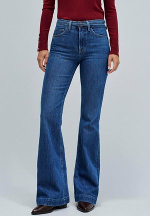 Vaqueros de mezclilla azul acampanados con cintura alta, cinco bolsillos y herrajes metálicos; textura suave y diseño estructurado, complementados con botas marrones.