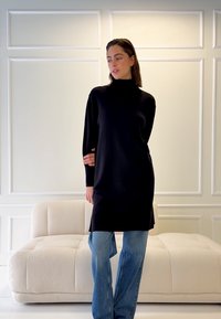 Svart poloklänning med lång ärm, avslappnad passform och små sidoskärningar, kombinerad med ljusblå jeans med rak ben på en mjuk beige soffa.