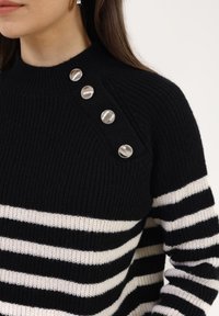 Femme portant un pull en tricot rayé noir et blanc avec quatre boutons argentés le long de la couture de l'épaule gauche.