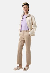 Krémová bunda s texturovaným límcem, světle fialová košile, béžové na míru šité kalhoty a bílé sandály s logem. Hladké textury po celém outfitu.