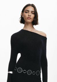 Maglione nero a spalla scoperta con texture a coste, caratterizzato da un accento argentato su una manica e una cintura circolare con borchie in vita.
