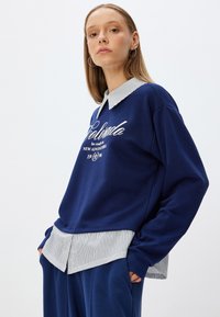Sudadera azul marino con texto en blanco, superpuesta a una camisa de cuello a rayas en azul claro y blanco, con mangas largas y un corte relajado.