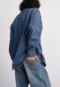 Denimskjorta med långa ärmar, blekt blå färg, synliga sömnadsdetaljer, avslappnad passform och en ficka med en läderlogotyp på baksidan.