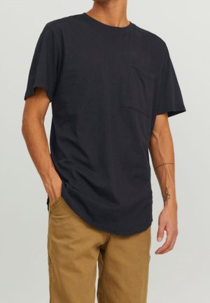 Homme portant un T-shirt noir uni à manches courtes avec une poche poitrine et un pantalon beige, debout avec une main dans une poche.