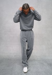 Grijze sweatshirt en sweatpants van zacht, gestructureerd stof. De outfit heeft een ontspannen pasvorm en subtiele logo borduursels op de broek, gecombineerd met witte sneakers.