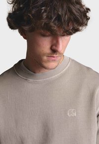 Sudadera beige claro con cuello redondo y puños acanalados, con un pequeño detalle de elefante bordado en el pecho. Textura suave.