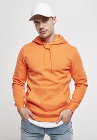 Sudadera con capucha naranja de tela suave, que presenta un bolsillo frontal, cordones ajustables y puños de canalé. Llevada con jeans de color azul claro.