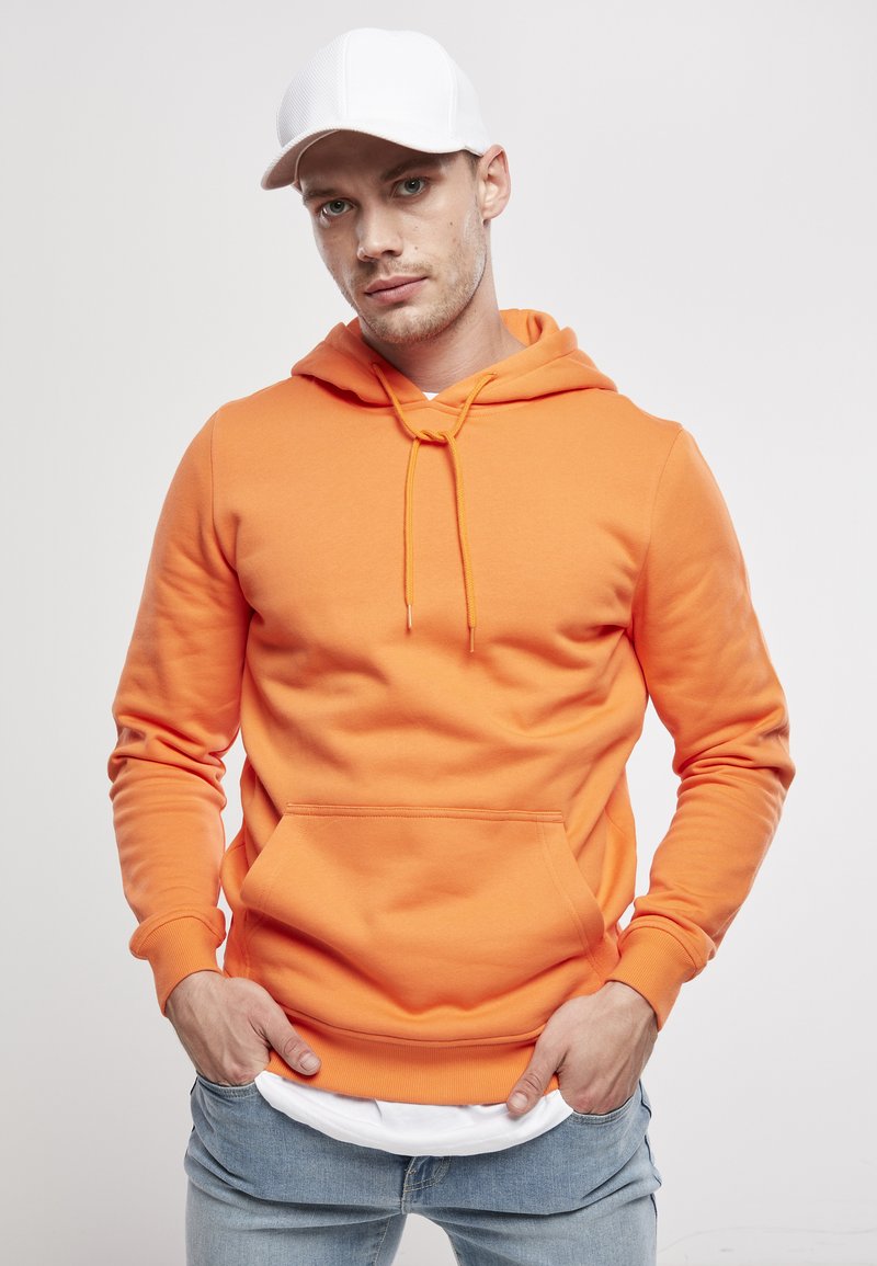 Sudadera con capucha naranja de tela suave, que presenta un bolsillo frontal, cordones ajustables y puños de canalé. Llevada con jeans de color azul claro.