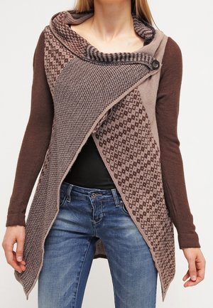 Kobieta w asymetrycznym, brązowym, dzianym kardiganie o fakturowanych wzorach, z zapiętym kołnierzem i niebieskich dżinsach, stojąca z luźno opuszczonymi rękami.