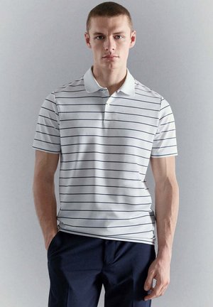 Joven con una camisa polo blanca con finas rayas negras horizontales y pantalones azul marino oscuro, de pie con una mano en el bolsillo frente a una pared gris.