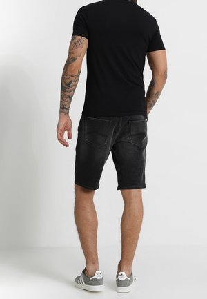 Mężczyzna w czarnej koszulce, czarnych dżinsowych szortach i szarych sneakersach Adidas stoi twarzą do białej ściany, prezentując wytatuowane ramiona.