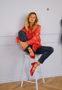 Frau mit langen blonden Haaren sitzt auf einem weißen Hocker vor einer hellen Wand, trägt eine rote Bluse mit Blumenmuster, dunkle Jeans und rote Absatzschuhe.