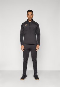 Chaqueta deportiva negra con cremallera y pantalones a juego, hechos de una tela suave; presenta un pequeño logo dorado en el pecho y zapatillas negras con suelas de goma.