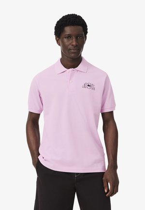 Hombre con camiseta polo Lacoste color púrpura claro con el logo en el pecho izquierdo y pantalones negros, de pie con las manos en los bolsillos frente a un fondo blanco.