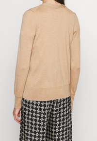 Beige tröja med långa ärmar, ribbade mudd och nederkant, tillverkad av fint stickat material. Bärs över en mönstrad svart och vit kjol.
