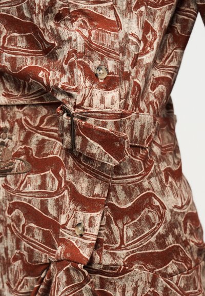 Κοντινό πλάνο καφέ και μπεζ υφάσματος με μοτίβα αλόγων, κλείσιμο με κουμπιά και υφασμάτινη ζώνη με λεπτομέρεια αγκράφας.