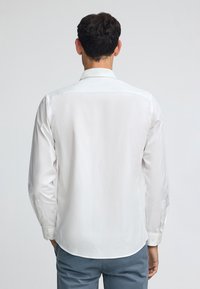 Vit långärmad skjorta med klassisk krage, med knappmanschetter och en slät textur. Bärs med ljusblå byxor.