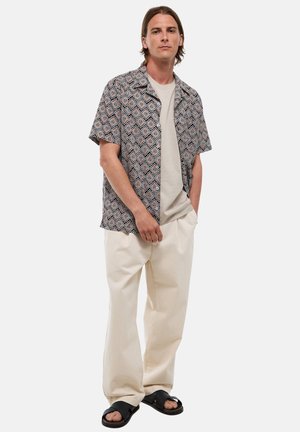 Homme portant une chemise à manches courtes à motifs par-dessus un T-shirt beige, pantalon ample de couleur crème et sandales noires, debout les mains dans les poches.