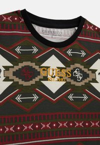 Organická bavlněná tričko se geometrickými vzory v tmavě zelené, červené a krémové barvě, s vyšitým nápisem "GUESS EST. 1981" v žluté. Černý výstřih.
