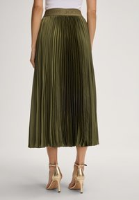 Woman wearing an olive green pleated midi skirt with a branded waistband and gold high-heeled ankle strap sandals, standing facing away.