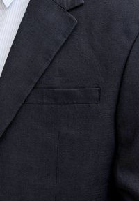 Tmavě modrý klopový límec saka a náprsní kapsa přes světle modrý a bílý proužkovaný límec košile, žádné viditelné doplňky ani značky.