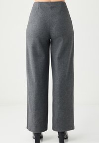 Frau trägt dunkelgraue, hochtaillierte, weit geschnittene Hosen und schwarze Absatzschuhe, von hinten vor weißem Hintergrund gezeigt.