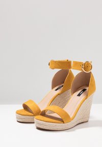 Žluté semišové klínové sandály s páskem kolem kotníku, které mají platformu obalenou jutou a textilní prvky. Design na nazouvání.
