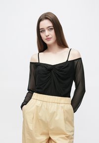 Svart axelbandslös topp med knutdetalj framtill och genomskinliga långa ärmar, kombinerad med höga ljusbeige byxor.