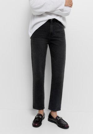 Persoon die zwarte rechte jeans draagt, een wit top met lange mouwen en zwarte leren instap loafers, staand tegen een effen achtergrond.