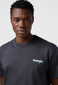 Tamsiai pilka trumpomis rankovėmis medvilninė marškinėliai su šviesiai mėlynu „Wrangler“ logotipu kairėje krūtinės pusėje. Lygios tekstūros, klasikinio stiliaus.