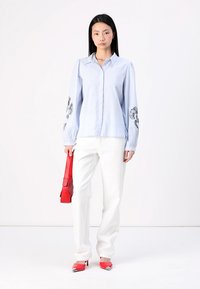 Camisa azul clara com mangas franzidas e bordado floral preto. Acompanhada de calças brancas e acessórios vermelhos, incluindo uma bolsa e sapatos.