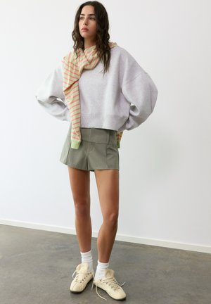Jonge vrouw met lang donker haar, draagt een grijze oversized trui, groene shorts, gestreepte sjaal, witte sokken en beige sneakers, staand binnenshuis.
