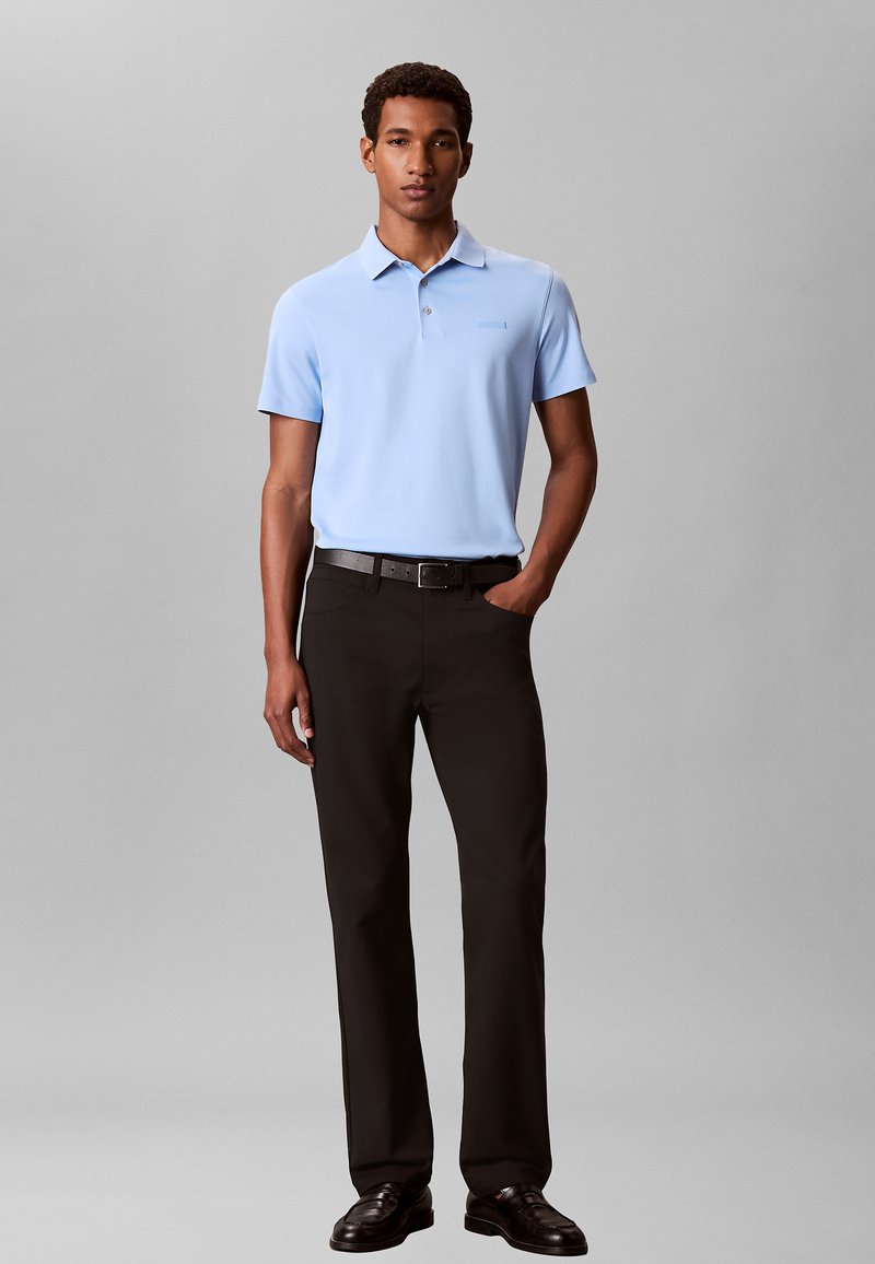 Man staat met één hand in zijn zak, draagt een lichtblauw poloshirt met korte mouwen, zwarte broek, zwarte riem en donkerbruine schoenen, grijze achtergrond.
