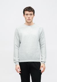 Világosszürke pulóver puha anyagból, bordázott mandzsettákkal és szegéllyel. Kerek nyakkivágással és raglán ujjakkal, fekete nadrággal párosítva.